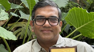 Photo of Jyotish Gopinathan holding the framed award citation with plants behind him.
