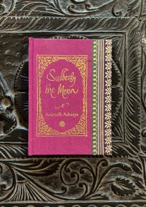 A sample cover of the book: bright pink handloom sari with gold embossed nameplate and lettering for the title and author's name. The border of the sari lines the right hand margin of the cover. The pocket book is placed against a black engraved background.