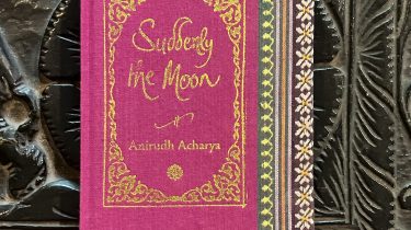 A sample cover of the book: bright pink handloom sari with gold embossed nameplate and lettering for the title and author's name. The border of the sari lines the right hand margin of the cover. The pocket book is placed against a black engraved background.